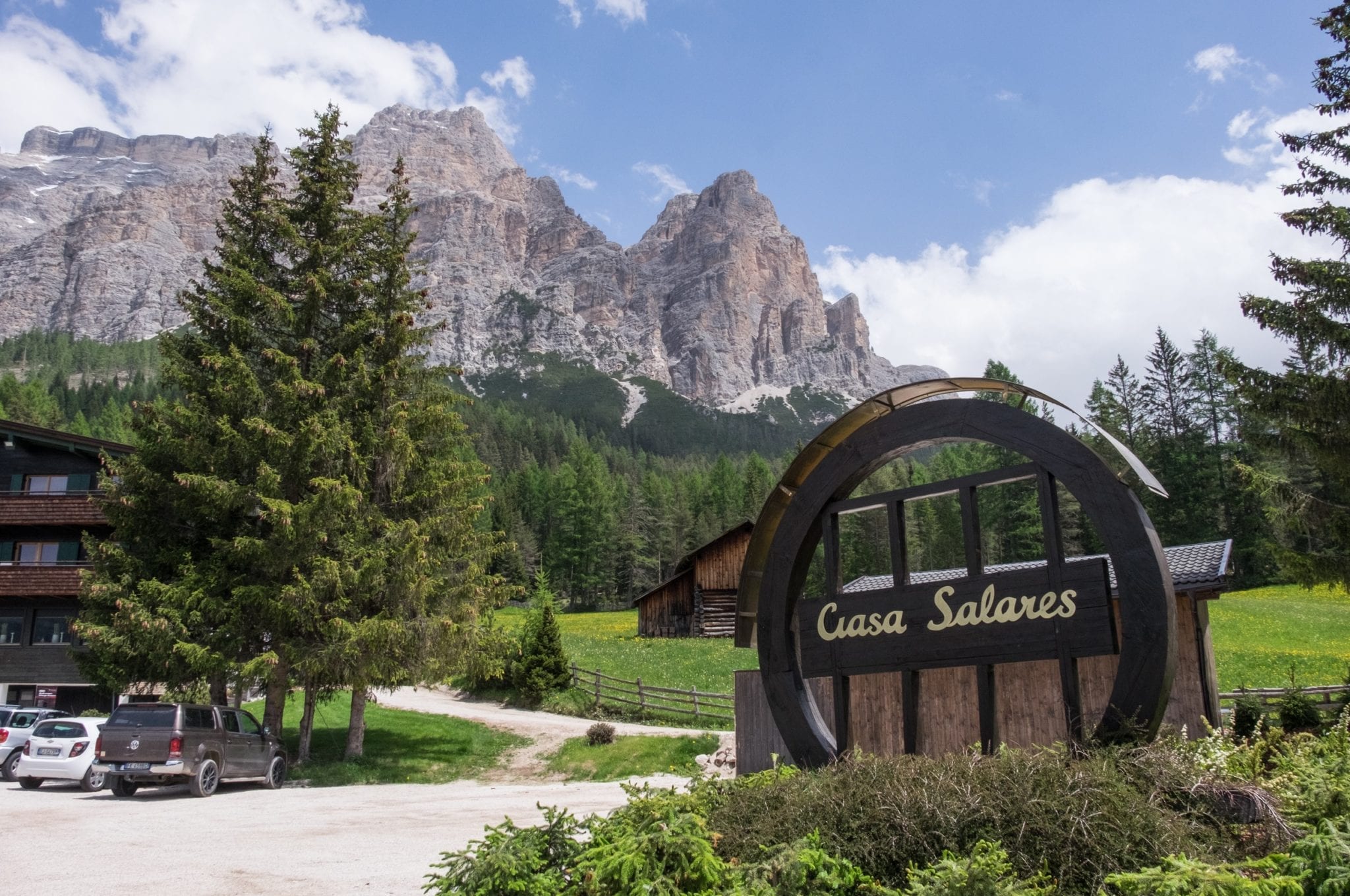 "Ciasa Salares" is written in a round sign in front of a backdrop of jagged gray mountains, green pine trees, and blue sky.