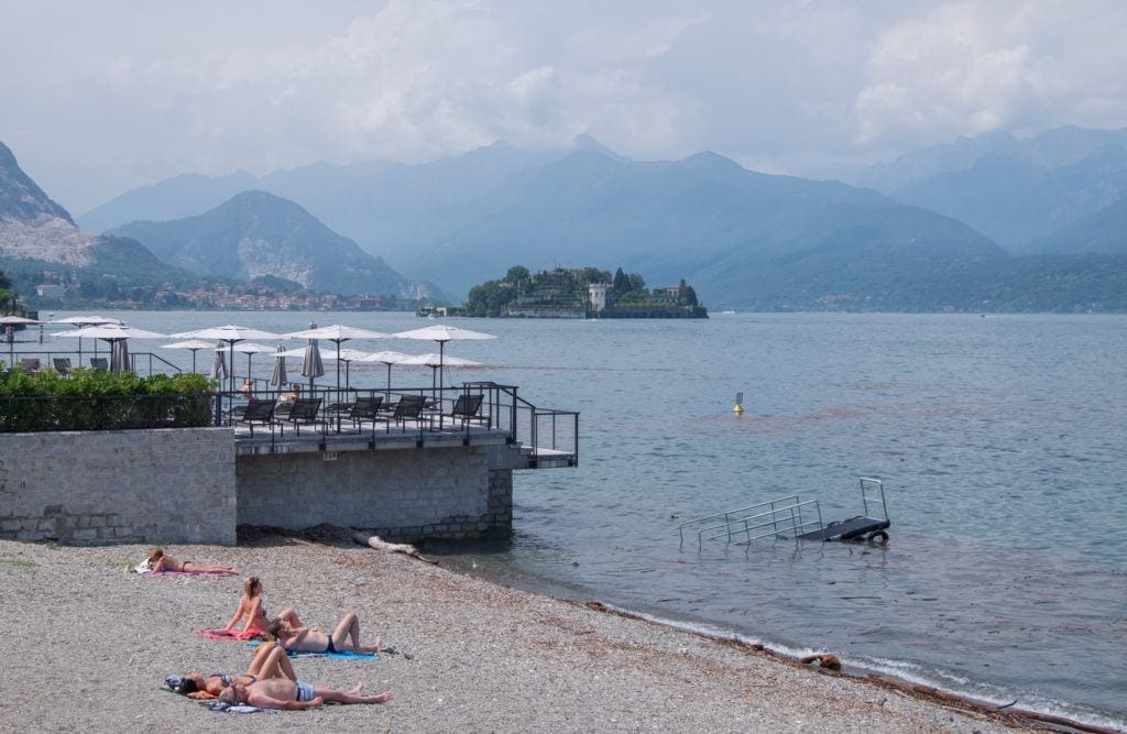 A man and a woman sunbathe on a gray rocky beach overlooking Lake Maggiore, which has an island in the distance and mountains rising up behind it underneath a cloudy sky.