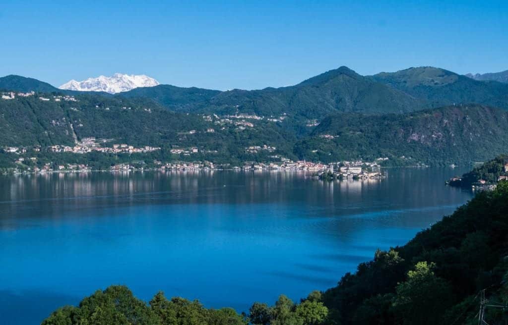 A bright blue Lake Orta with a tiny island in the lake, green mountains on the other side, and a glacier-covered white mountains in the distance, all underneath a bright blue sky.