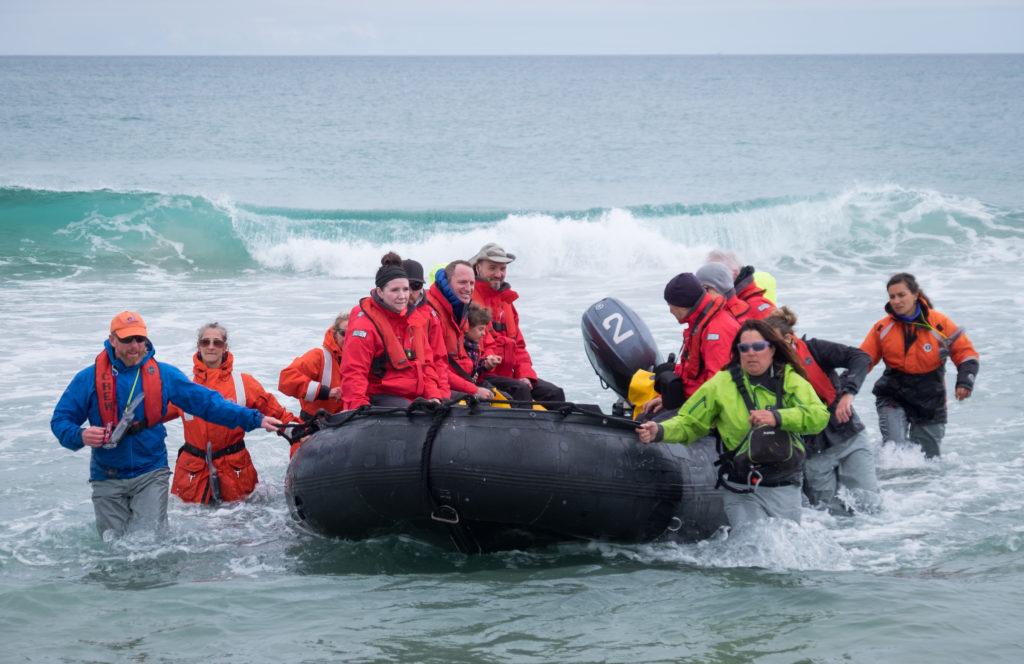 A zodiac full of people in red jackets is caught by several crew members in a gray, violent surf.