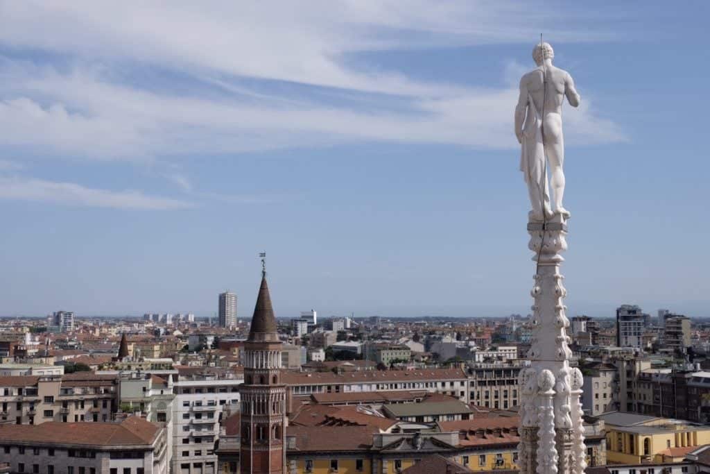 View from Milan's Duomo: A nude statue looks over the city from behind; the city is a mix of old Renaissance towers and modern buildings underneath a periwinkle blue and white striped sky.