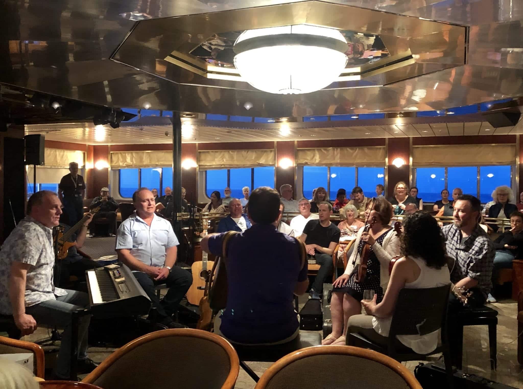 The Barra MacNeills band plays with the members sitting in a circle with violins, keyboard, and drums, surrounded by passengers on the cruise ship.