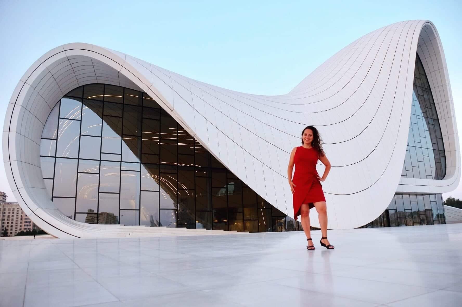 Kate stands in front of a modern building shaped like a sideways S, standing in a red dress.