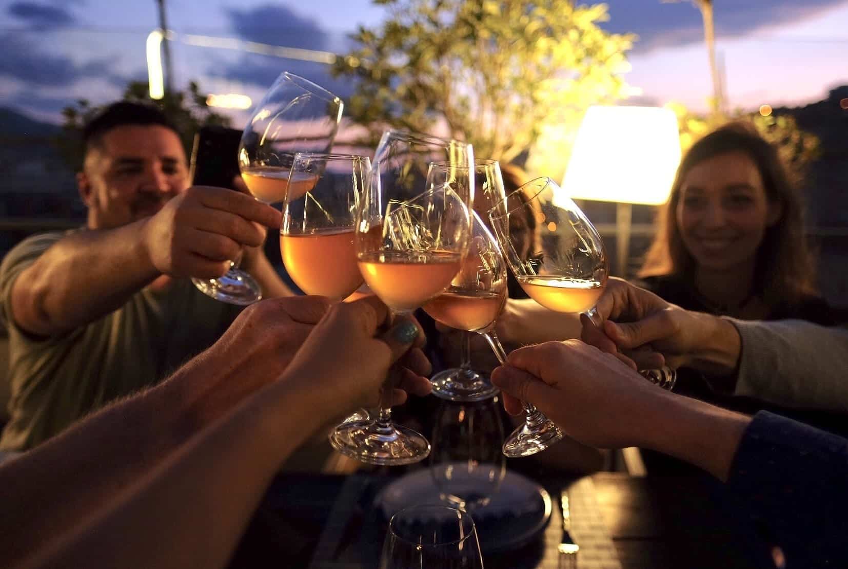 Several people toasting glasses of orange wine on a darkened rooftop at sunset.