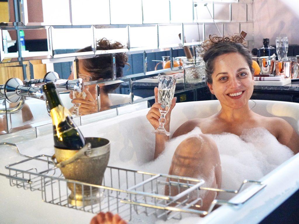 Kate in a bubble bath holding a glass of champagne with her hair up and smiling.