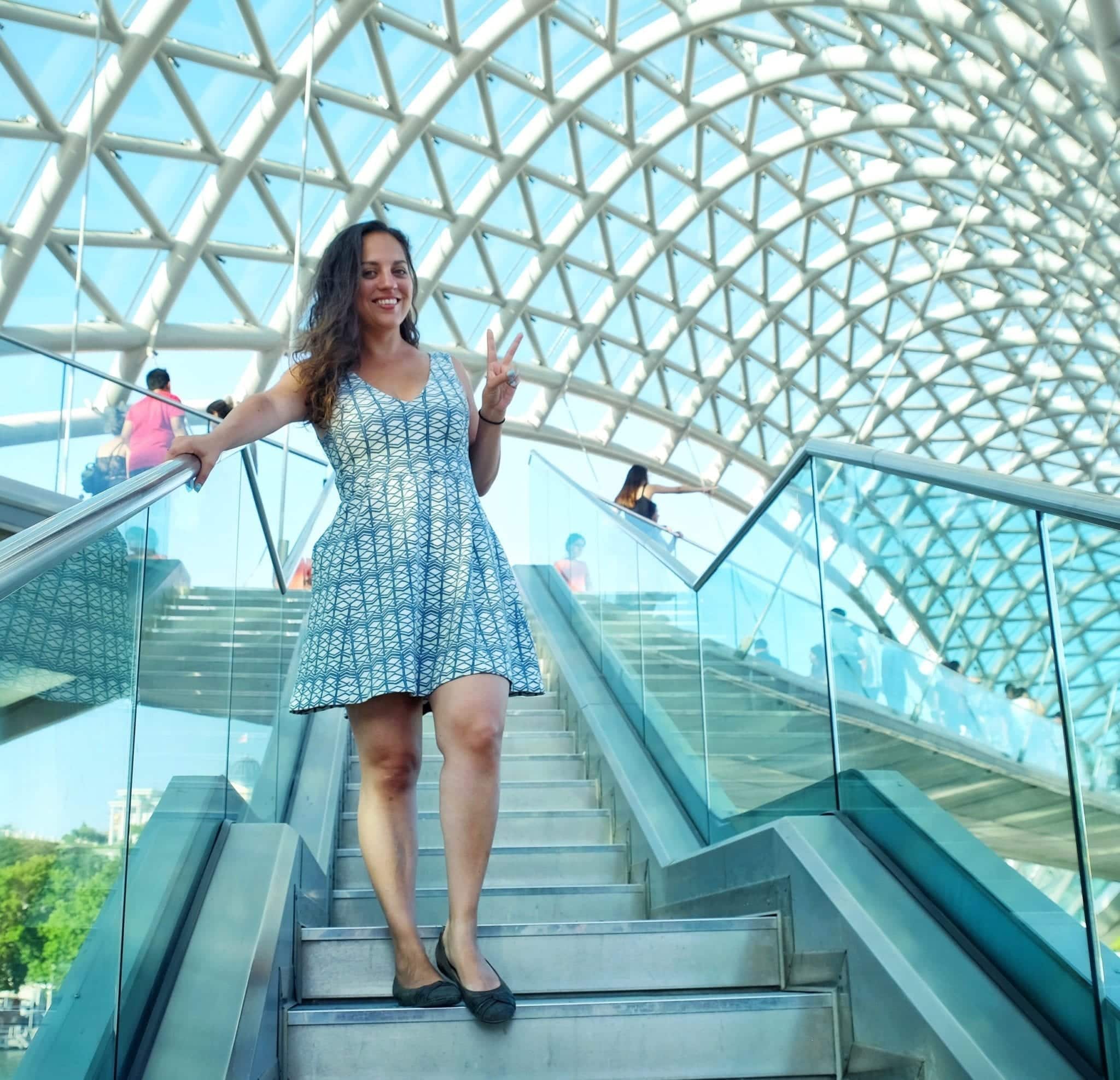 Kate stands underneath the green-and-white-patterned Peace Bridge in Tbilisi, Georgia, giving a peace sign and wearing a dress that has a very similar green and white geometric pattern.