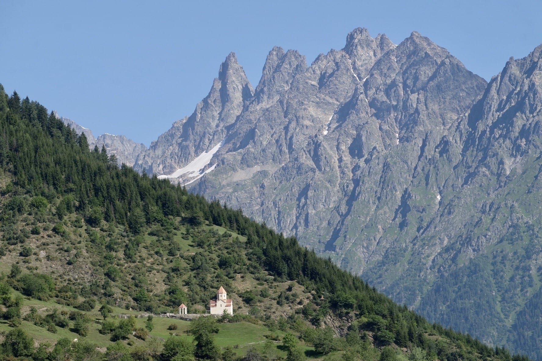 Two mountains: a green one with a tiny white church perched on it, in front of a taller, jagged gray mountain in Svaneti, Georgia.
