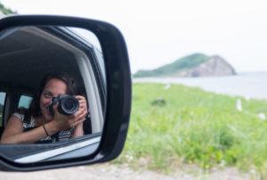 In the rearview mirror, you see Kate taking a photo with her DSLR camera. In the distance is a rock formation in the ocean.