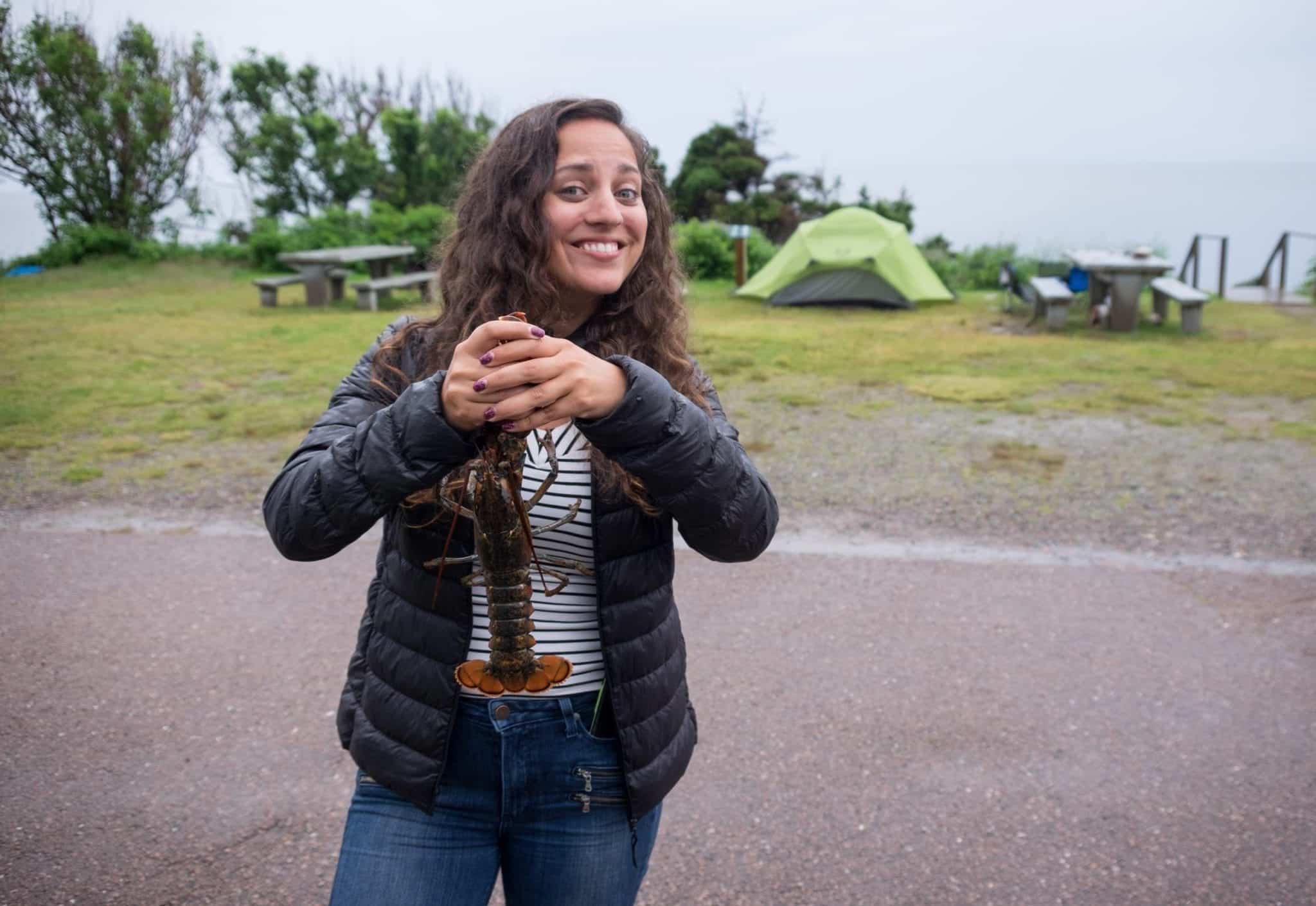 Kate holds a live lobster by the claws and smiles nervously. Tents are behind her.