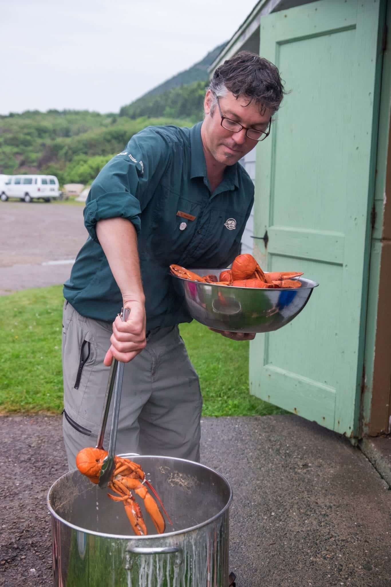 Parks Canada guide Robert holds a bowl of cooked lobsters in one hand and fishes them out of the pot with tongs.