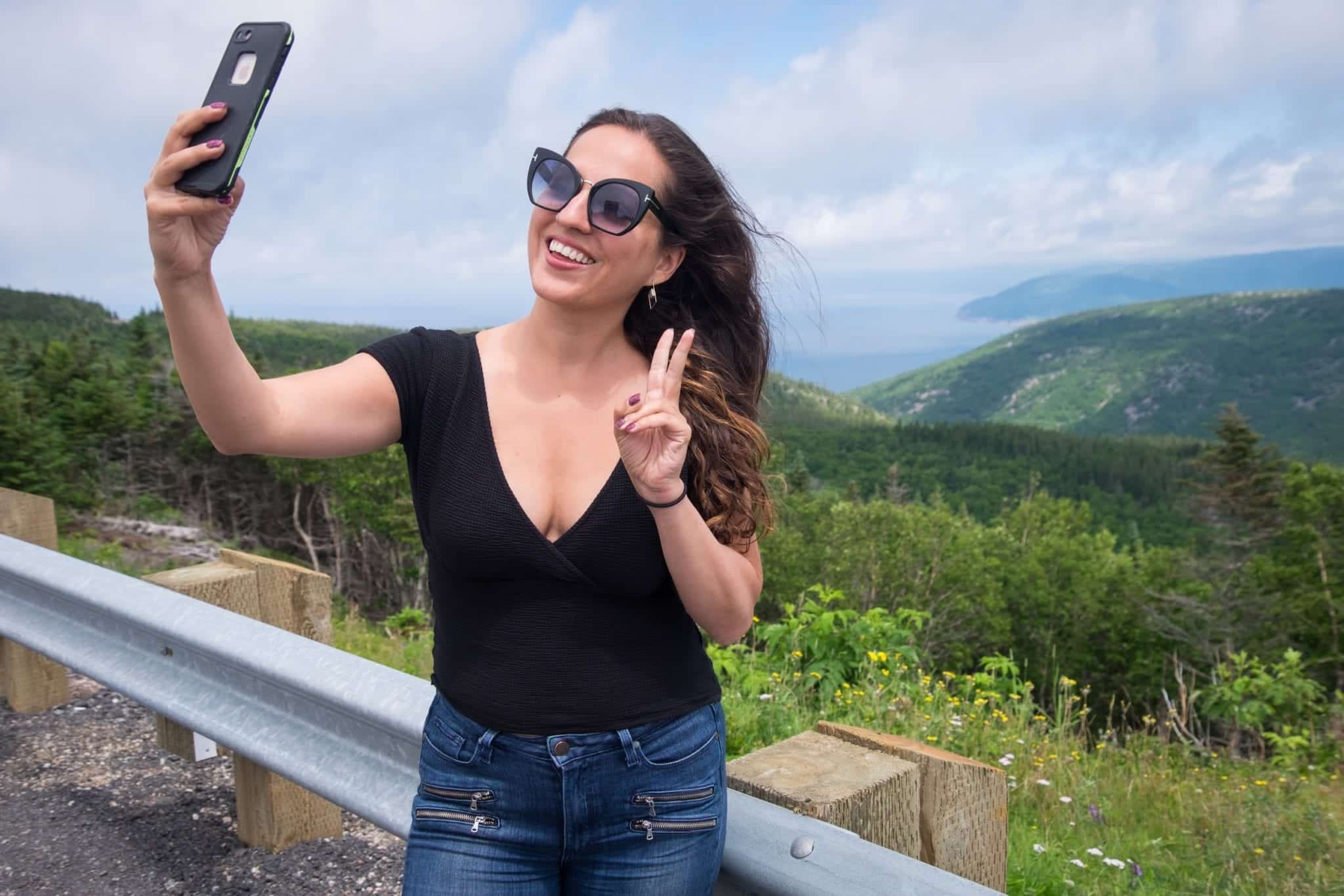 Kate wears large sunglasses and takes a selfie in front of hills and oceans at Cape Breton Highlands National Park