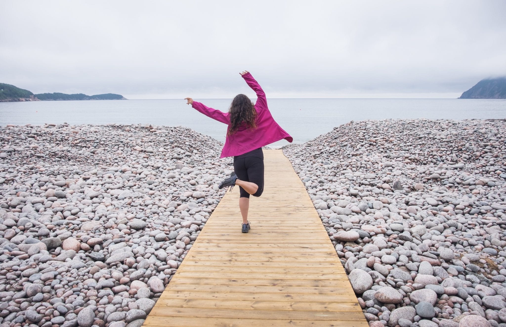 Kate wears a hot pink coat and dances on a platform at Ingonish Beach, surrounded by rocks.