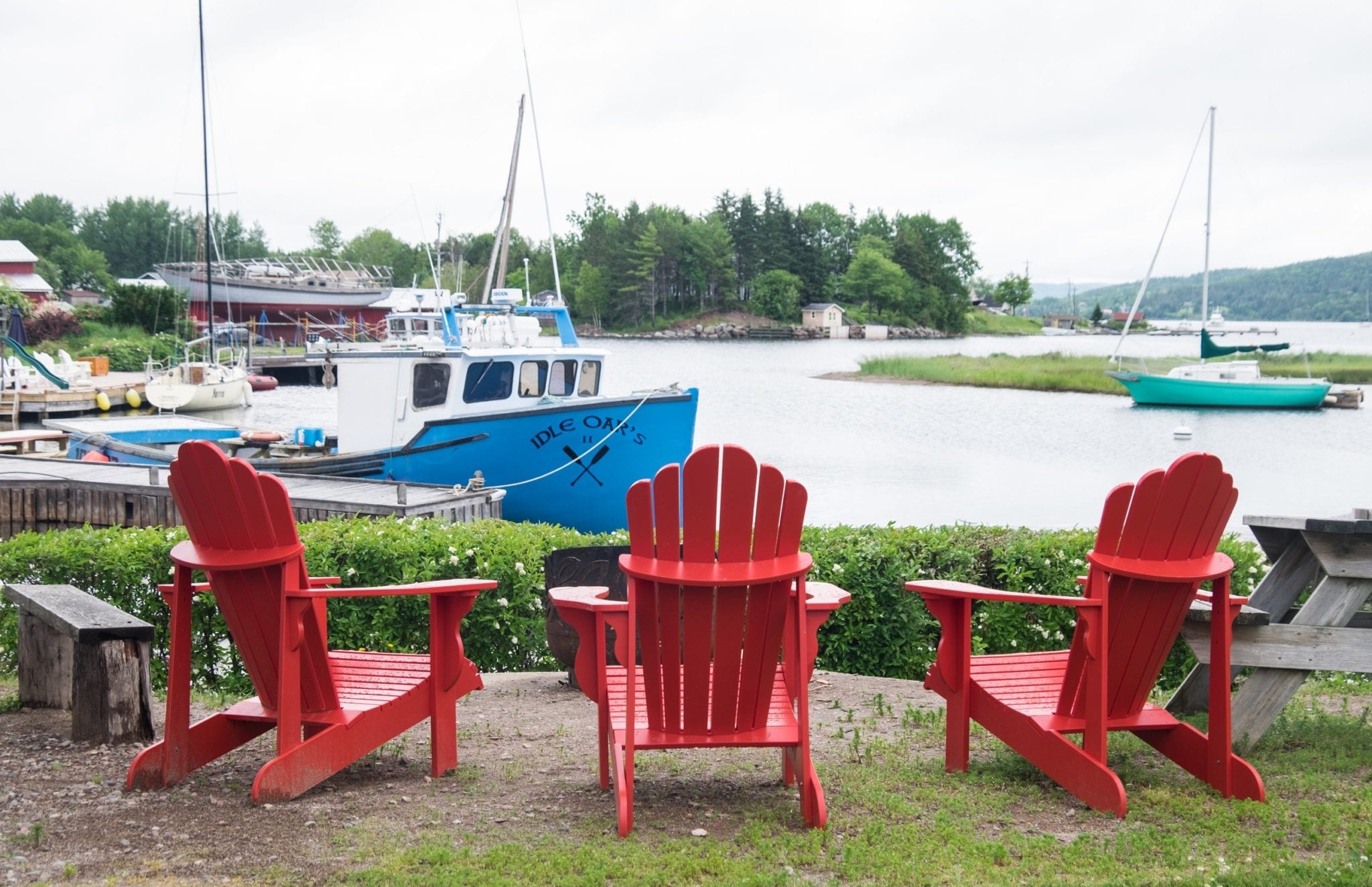 Three red chairs sitting in front of a blue boat a lake on an overcast day in Nova Scotia.