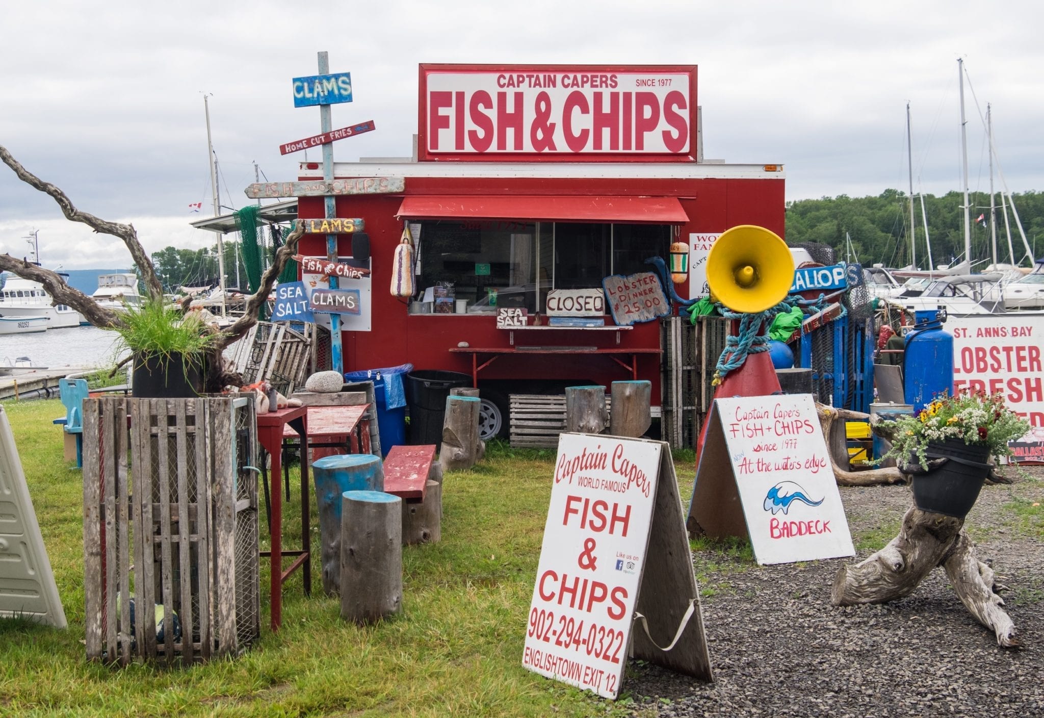 A red fish and chips food stand in front boats in Baddeck, Novia Scotia with signs saying "Fish and Chips" and tree stumps to sit on