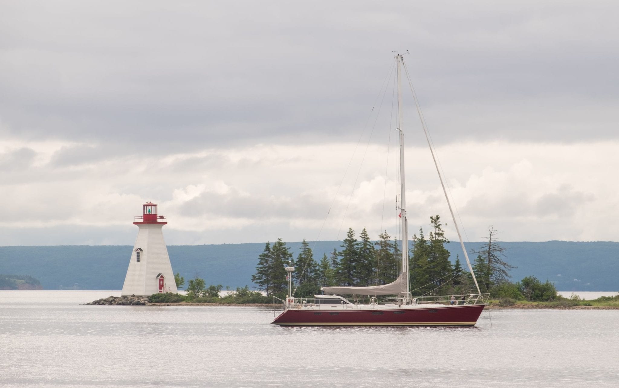 A white lighthouse with a red top on a tiny island filled with pine trees in a calm bay. In the foreground is a sailboat.
