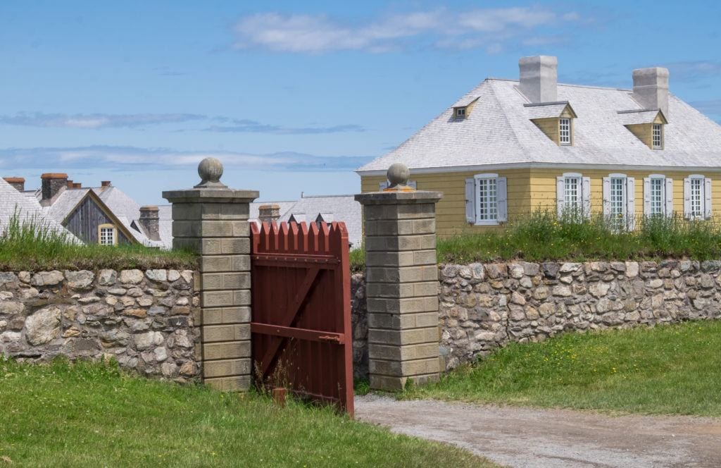 A stone wall has a red wooden gate open. There is a yellow building in the background underneath a blue sky streaked with white clouds. Louisberg, Nova Scotia.