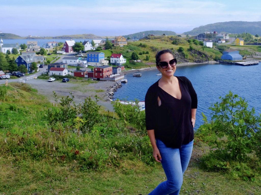 Kate stands in front of a panorama of Trinity, Newfoundland: green hills with brightly colored cottages next to the sea.