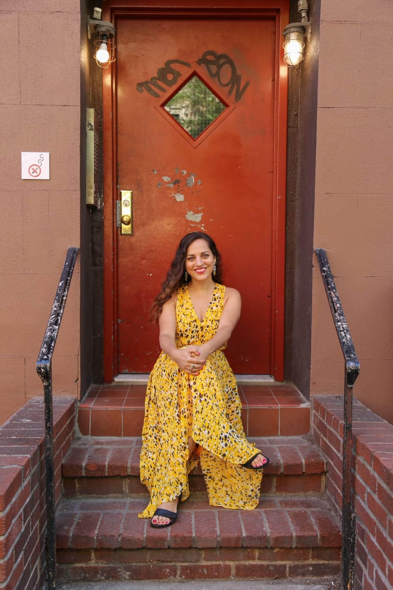 Kate sitting on a stoop in front of a red door with a diamond-shaped window in SoHo, NYC.