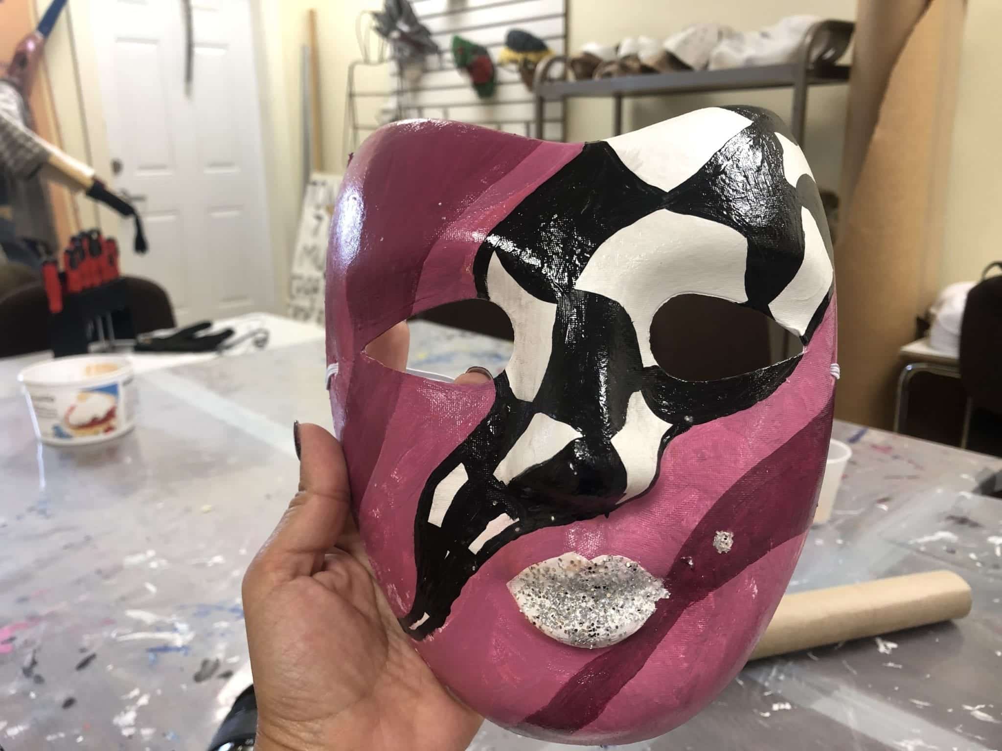 Kate holds her painted Mi-Careme mask -- pink and purple stripes with a black and white checked design running diagonally across the side, with silvery glitter lips.