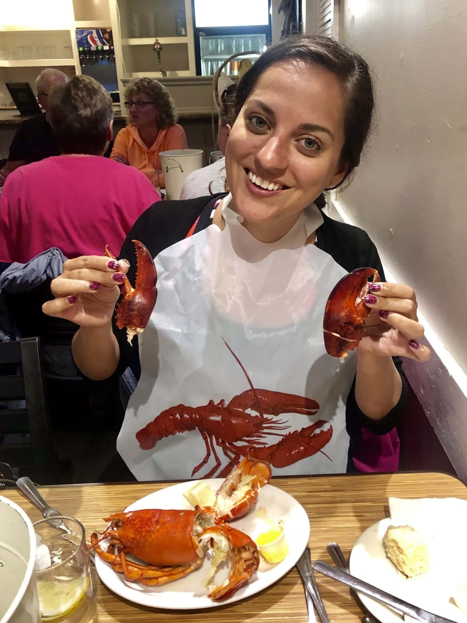 Kate smiles and holds up two lobster claws while wearing a lobster bib.