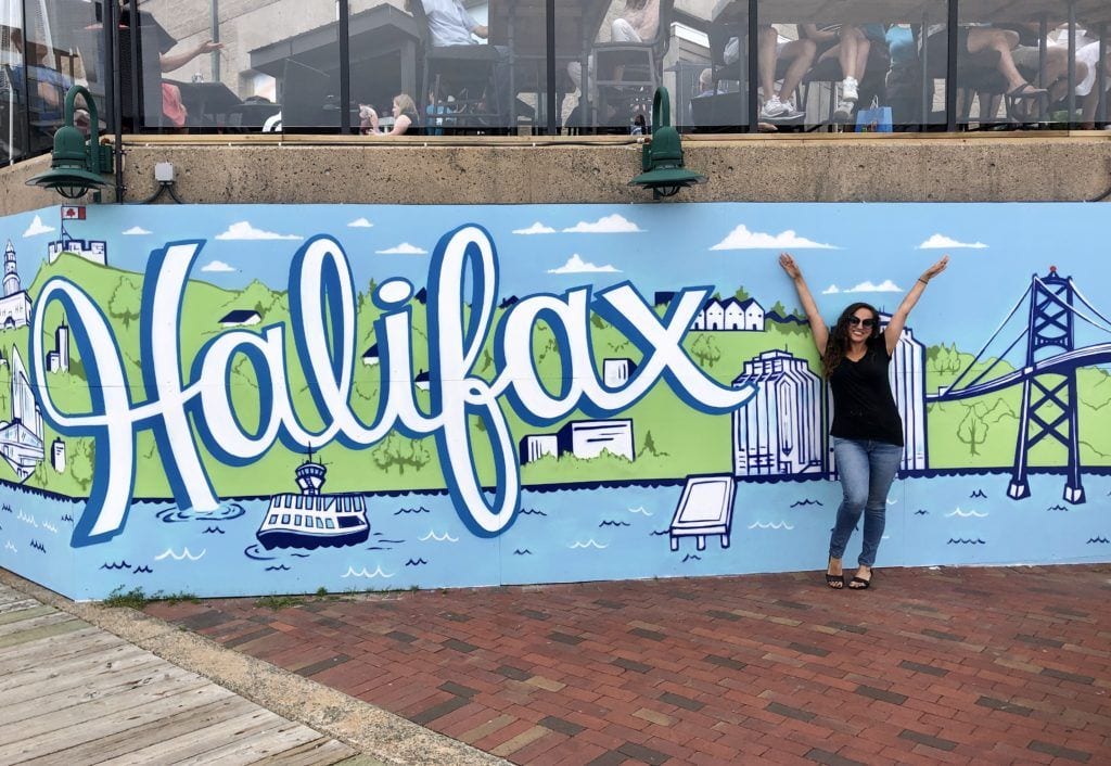 Kate poses with her arms in the air in front of a blue, green and white mural with the word Halifax in front of the Halifax cityscape.