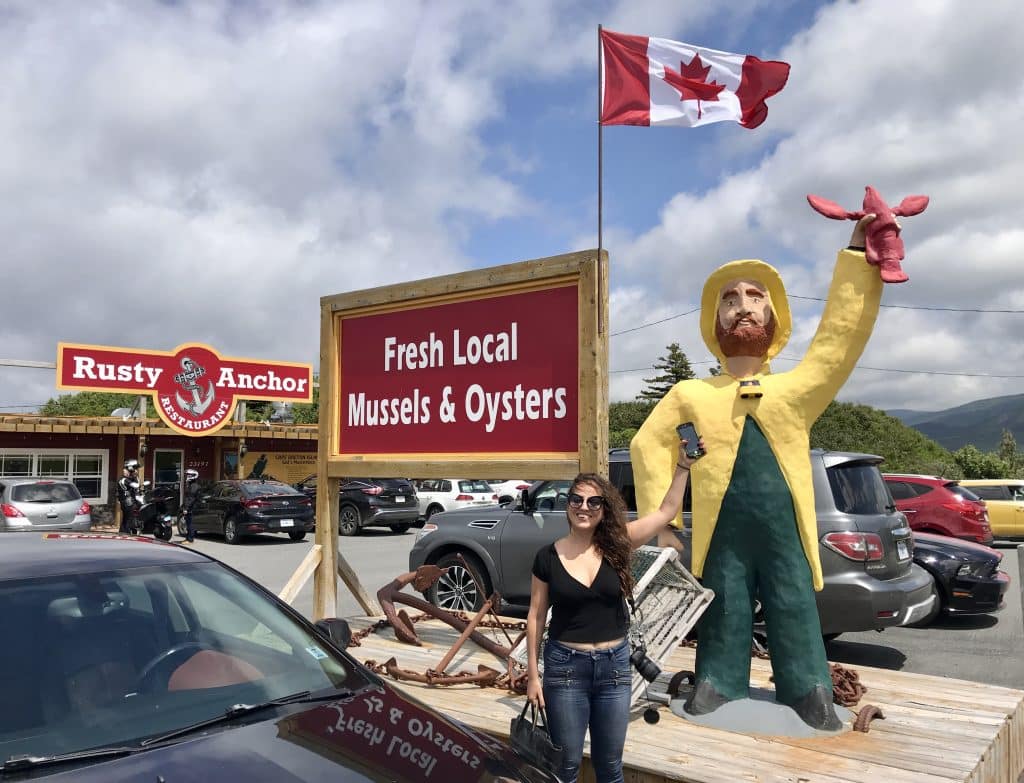 Kate standing and holding her phone up next to a statue of a fisherman holding a lobster up, a Canadian flag behind them.