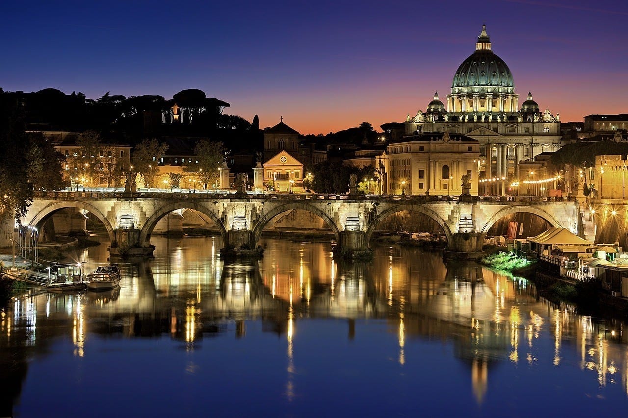 A purple and pink sunset in Rome. You see the lights of a bridge reflecting in the river, and on the right is the dome of St. Peter's Basilica.