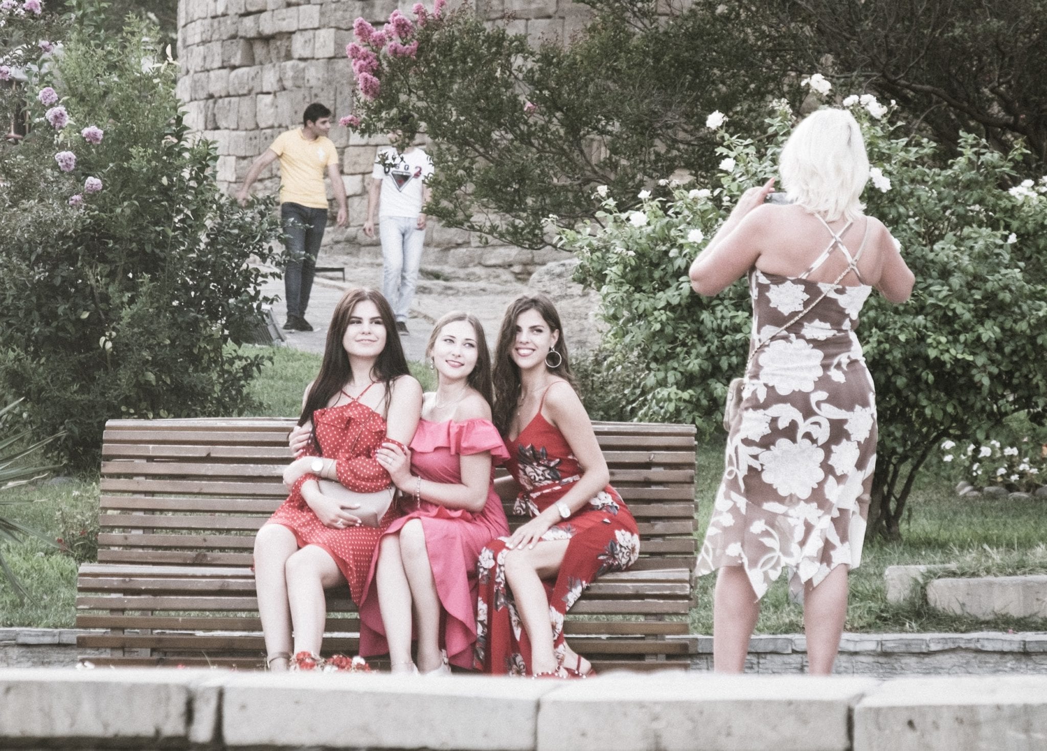 Tre ragazze adolescenti in abiti rosa e rossi sedute su una panchina nella città vecchia di Baku mentre una donna scatta la loro foto.