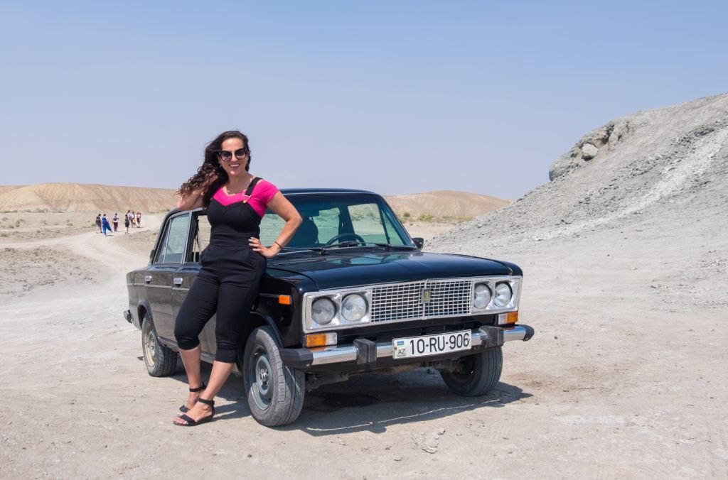Kate wears overalls and a pink shirt and poses with her hand behind her neck while sitting on a Soviet-era Russian Lada car. She's in the middle of the gray-brown dessert beneath a pale blue sky.
