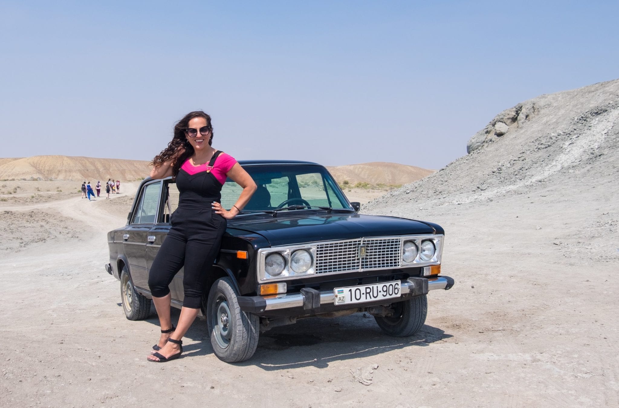  Kate porte une salopette et une chemise rose et pose la main derrière le cou assise sur une voiture Lada russe de l'ère soviétique. Elle est au milieu du dessert gris-brun sous un ciel bleu pâle.