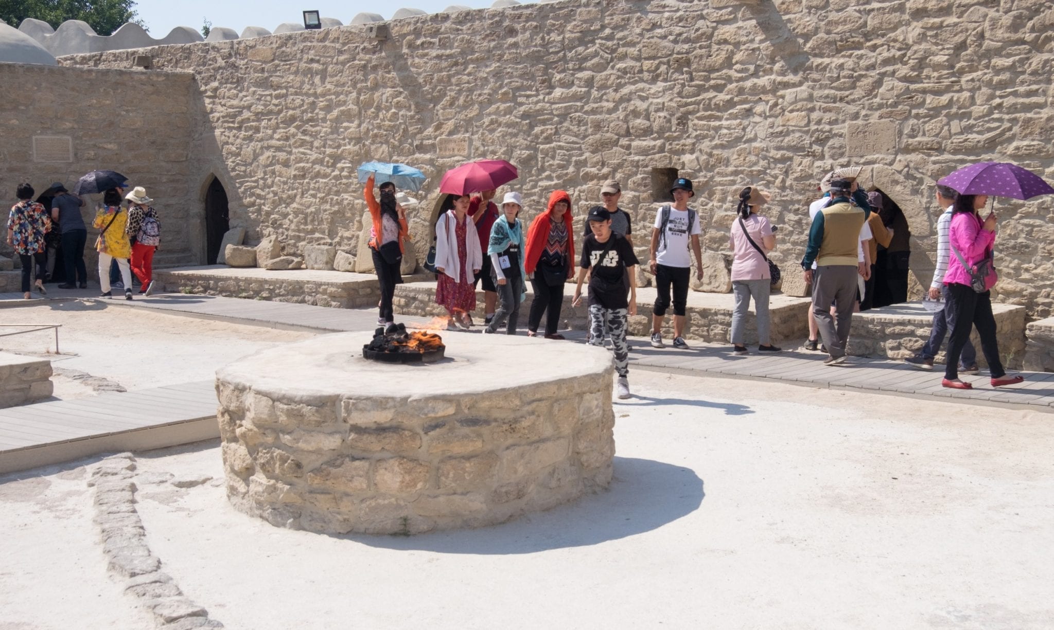 Fire bursts out of a well-like opening at Ateshgah Temple; behind it, tourists walk in a line and explore.