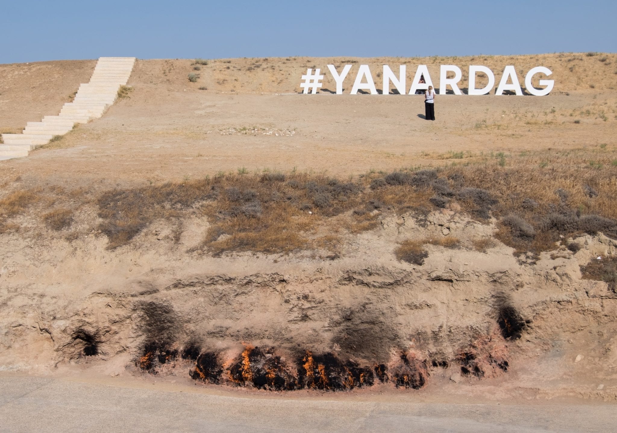 Ein brauner Hang bei Yanar Dag, wo unten Flammen aus der Erde ausbrachen. Es sagt #YANARDAG in Weiß auf dem Hügel, im Stil des Hollywood-Zeichens.