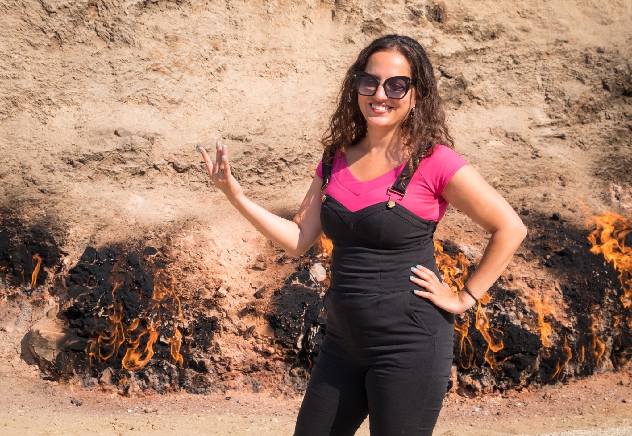 Kate stands in front of the burning ground at Yanar Dag in Azerbaijan, wearing overalls and holding her hand up and using her fingers to mimic flames.