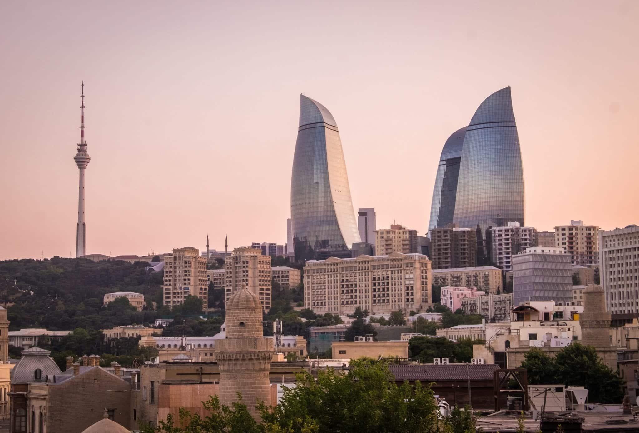 tři plamenné věže v Baku, Ázerbájdžán vypadají modro-fialové proti růžové obloze za soumraku. Pod nimi jsou menší budovy a vlevo je vysoká, hubená televizní věž ve městě.