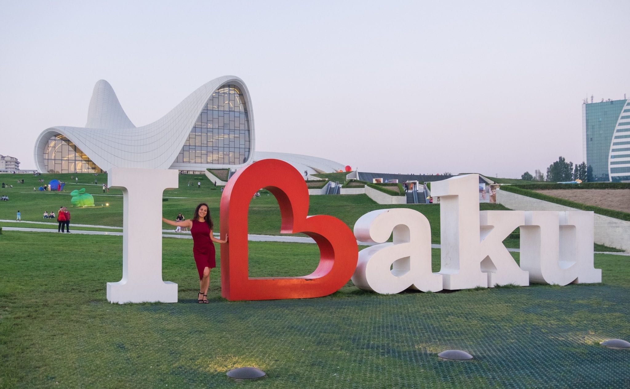 Kate stands in the middle of an "I Love Baku" sign where the B is shaped like a heart. In the background is the swooping white roof of the Heydar Aliyev Center.
