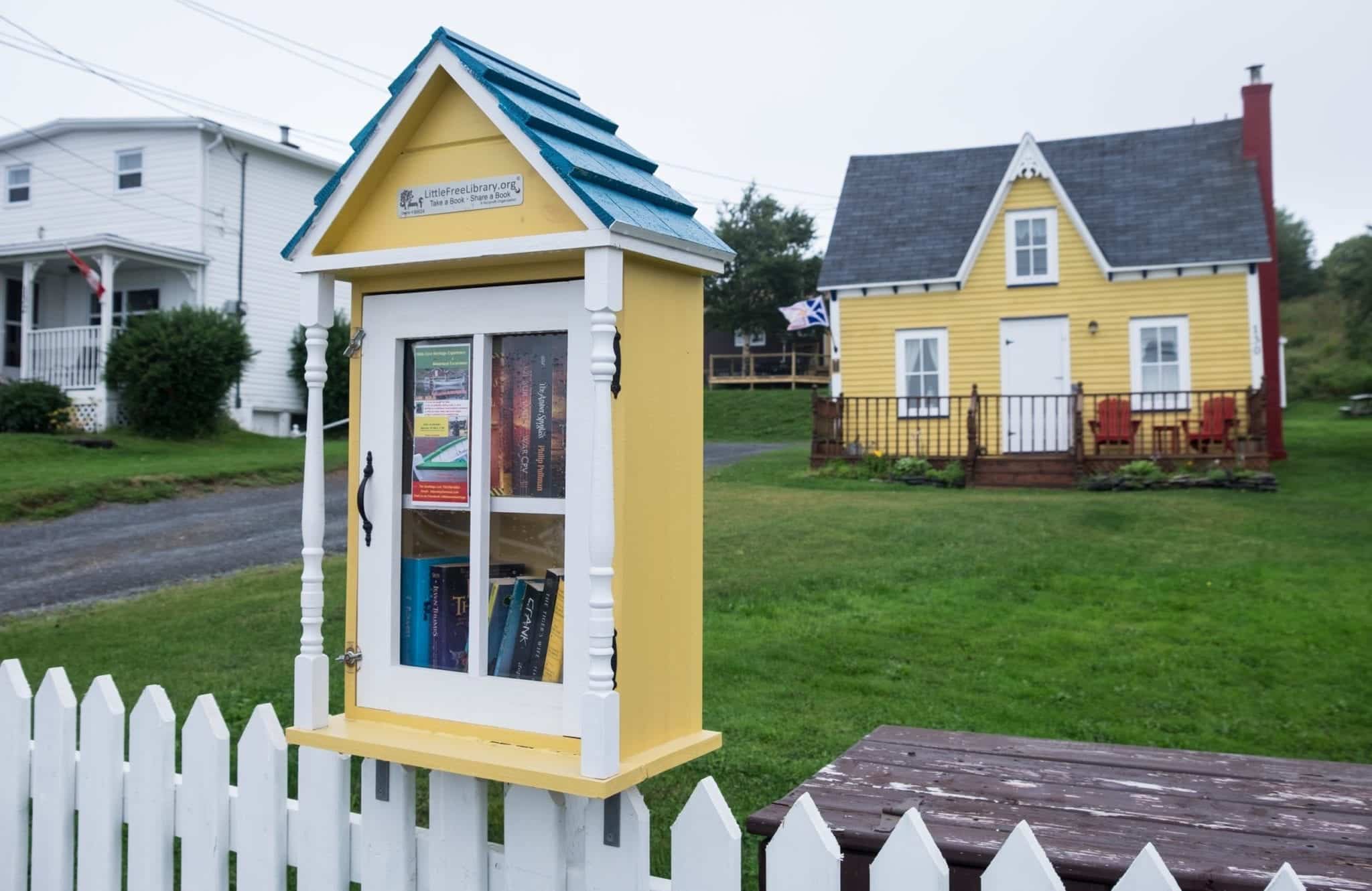 A yellow little library built on a white picket fence with books behind the glass door. In the background, a matching yellow cottage.