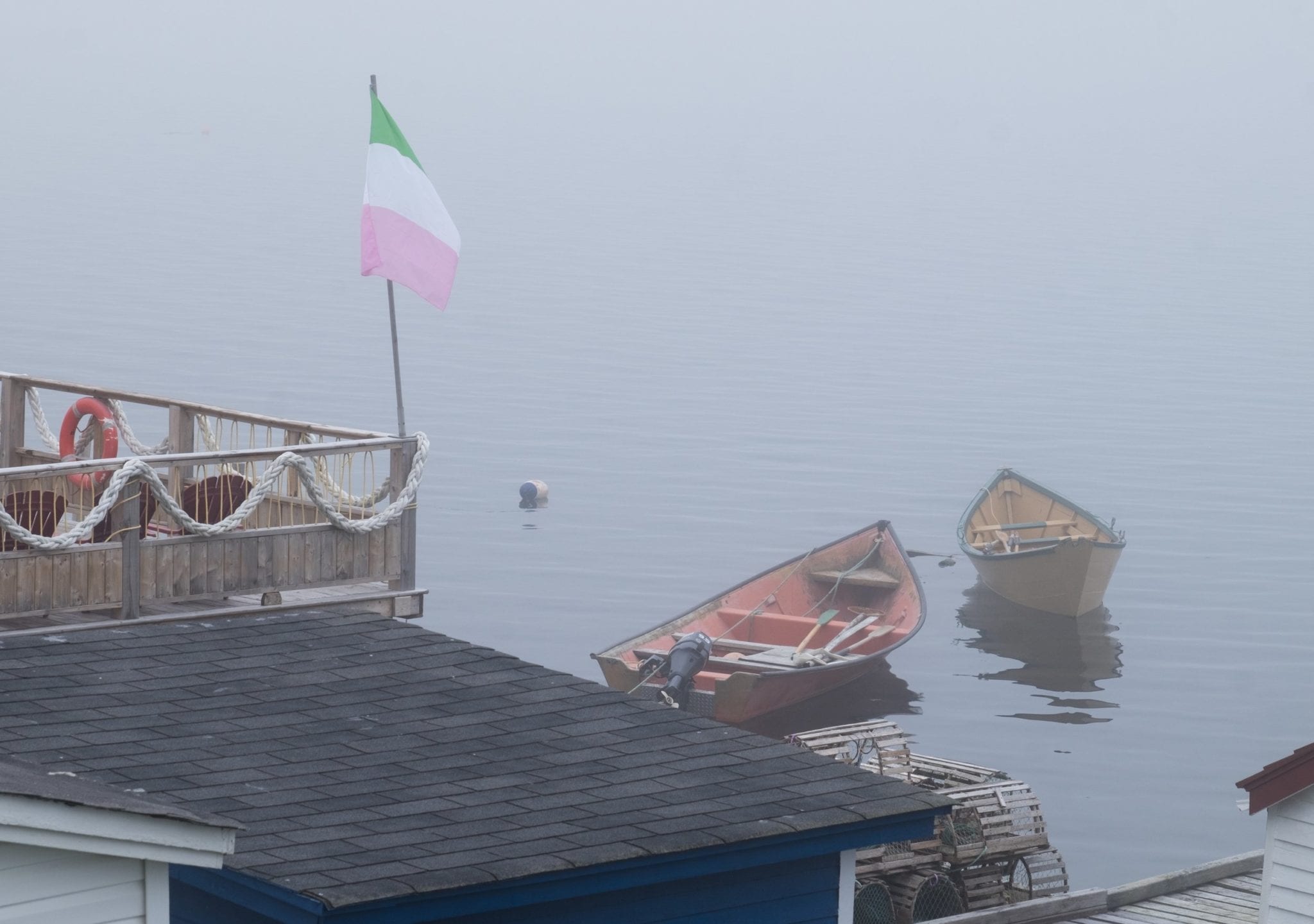 Rowboats in a foggy gray harbor. A flagpole with the green, white and pink Newfoundland independence flag on top.