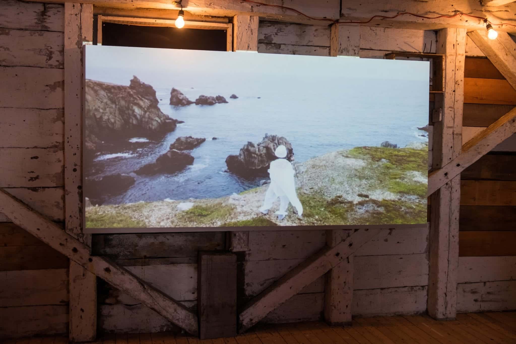 A screen with a woman in white African costume and a turban, standing on the edge of a cliff in Newfoundland.