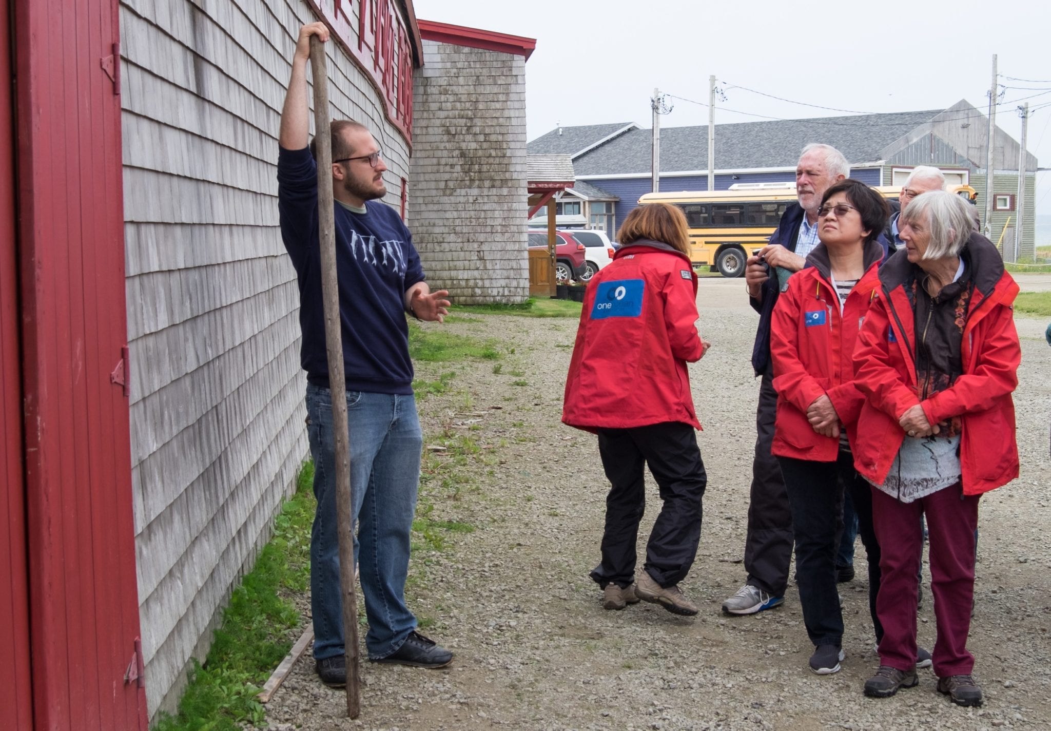 The smokehouse owner stands in front of the smokehouse holding a stick taller than him. Several older people wearing bright red OneOcean jackets listen to him talk.