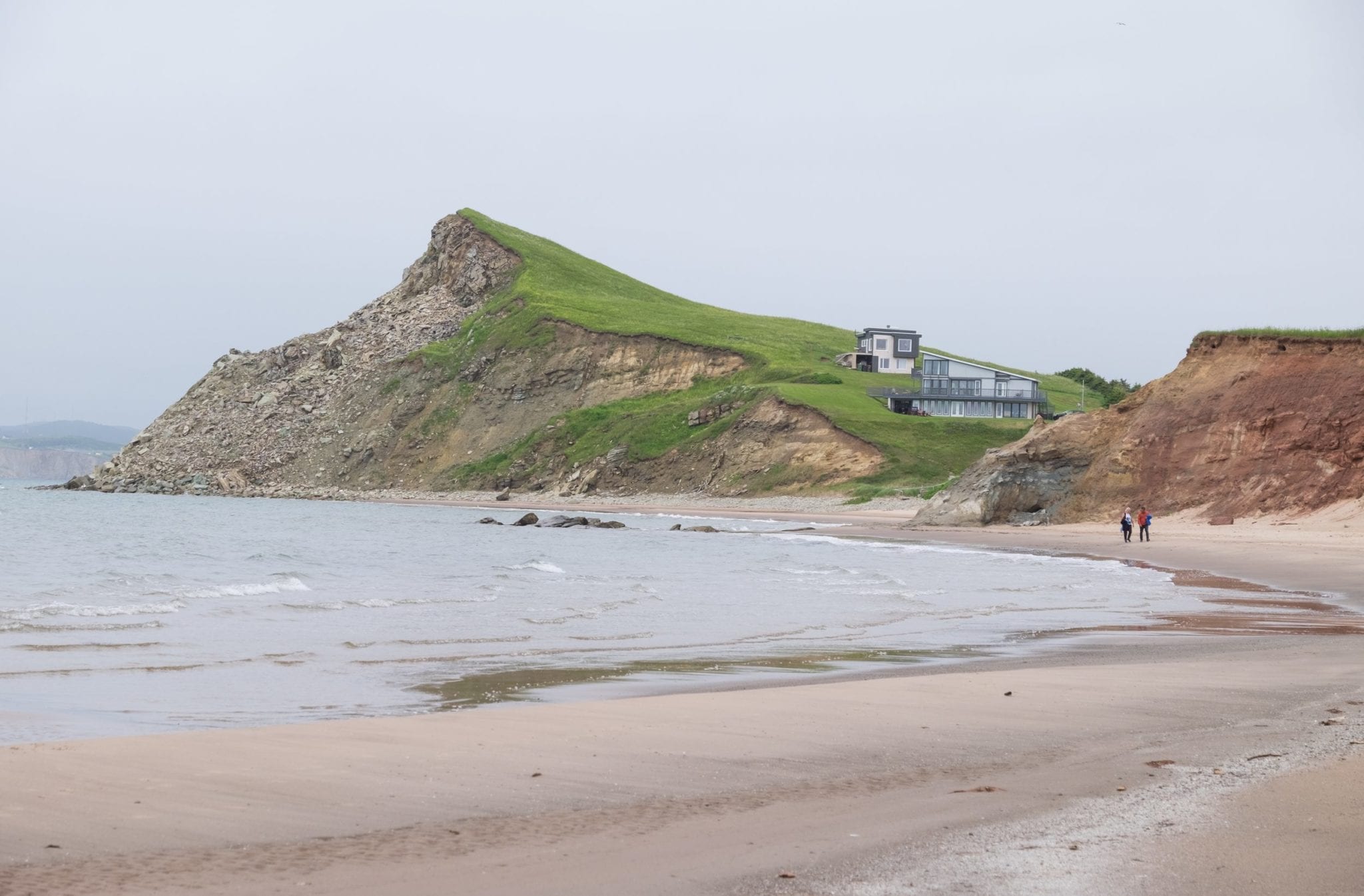 A gray-beige beach with a pointy green hill in the background.