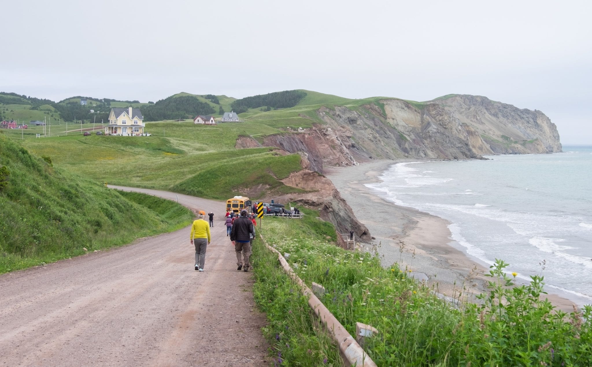 A group walks down a dirt road in the Iles-de-la-Madeleine; on the right is a gray cliff leading down to a rocky beach.