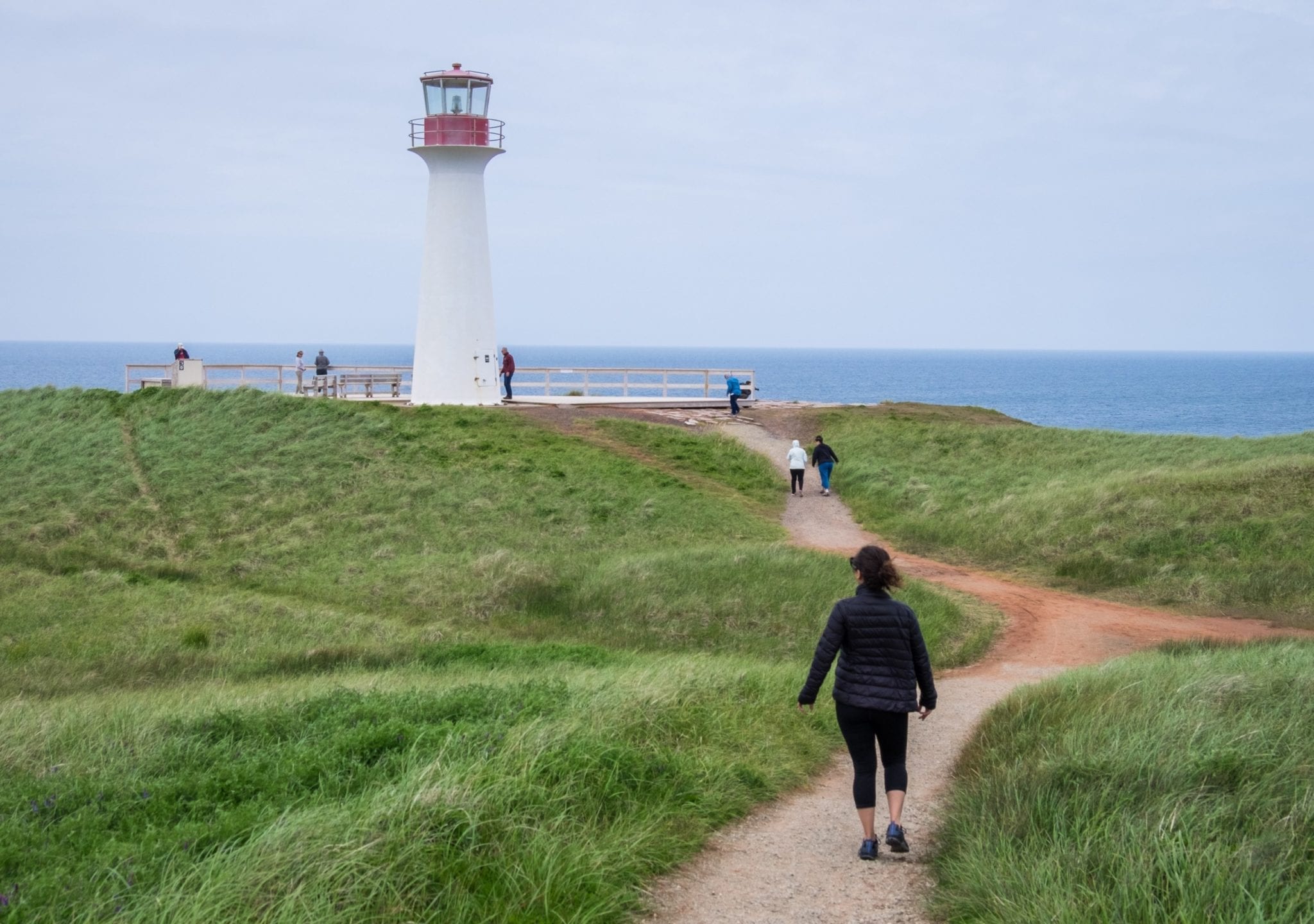 Kate walks along the path on a grassy hill to a white lighthouse in the Iles-de-la-Madeleine.