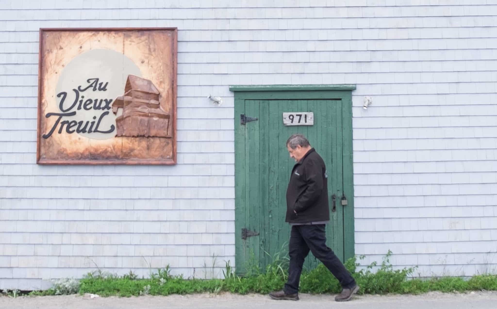 A gray house with a green door in La Grave as an older man walks by in front of the door, his head pointing down as if deep in thought.