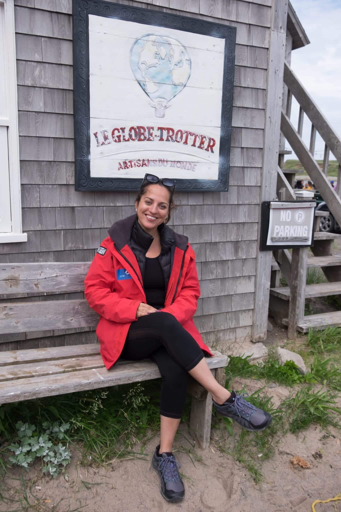 Kate wears a red waterproof coat over black leggings and sits on a bench in front of a gray house with a sign that reads "Le Globetrotter".