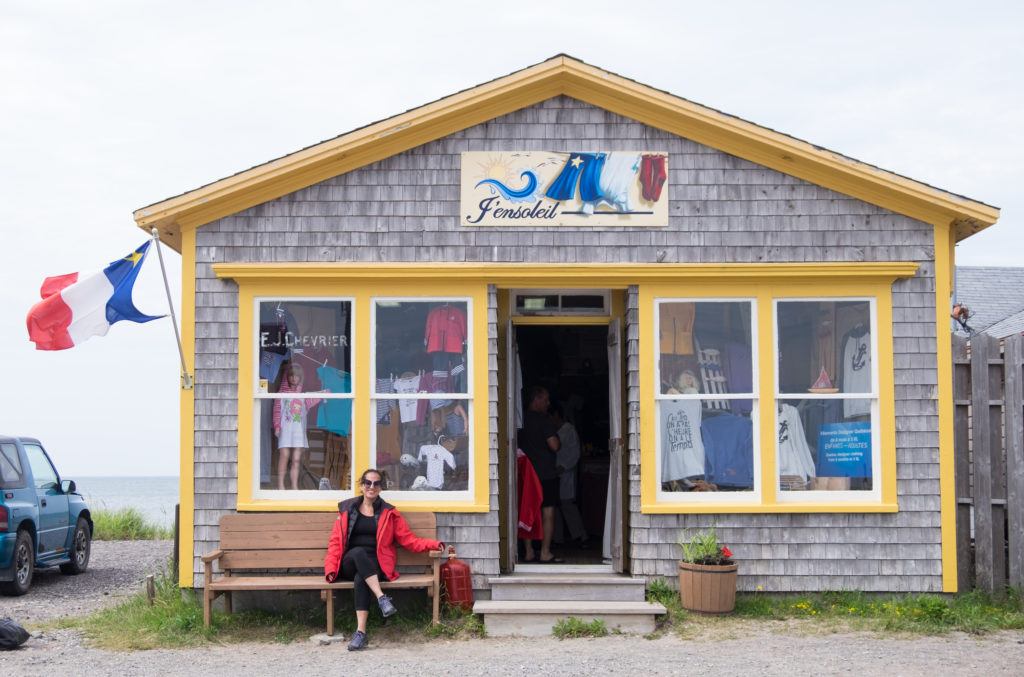 Kate sits on a bench in front of a gray shop with yellow trim and a red white and blue Acadian flag with a yellow star on it.