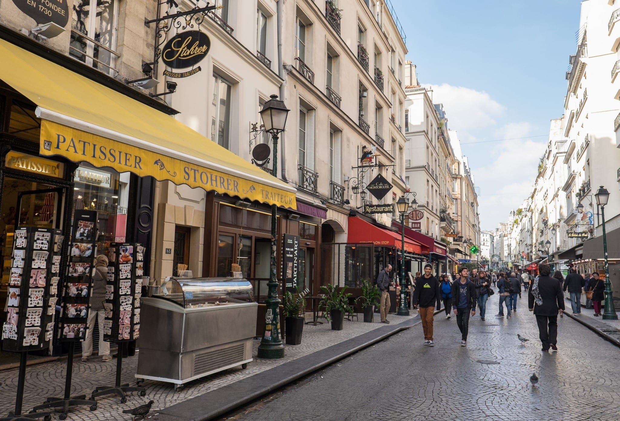 Best Places to Stay in Paris: a photo of Rue Montorgueil, lined with food shops and people walking down the street. In front is a shop with a bright yellow awning.