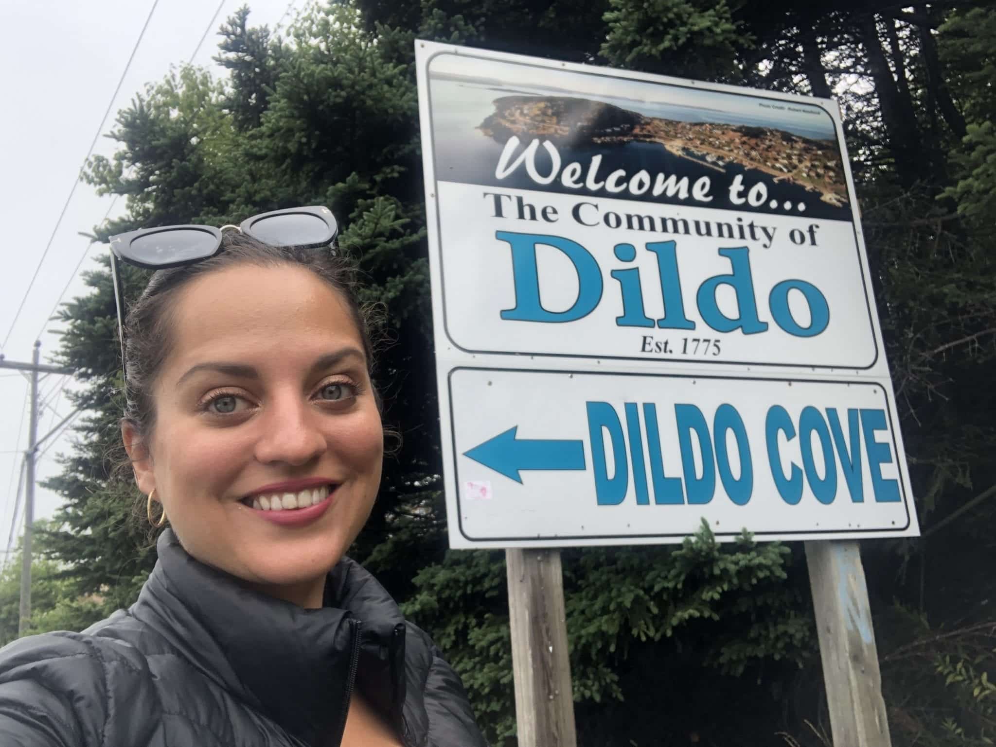 Kate stands next to a sign that reads "Welcome to the community of Dildo" and "Dildo Cove" with an arrow pointing to her face.