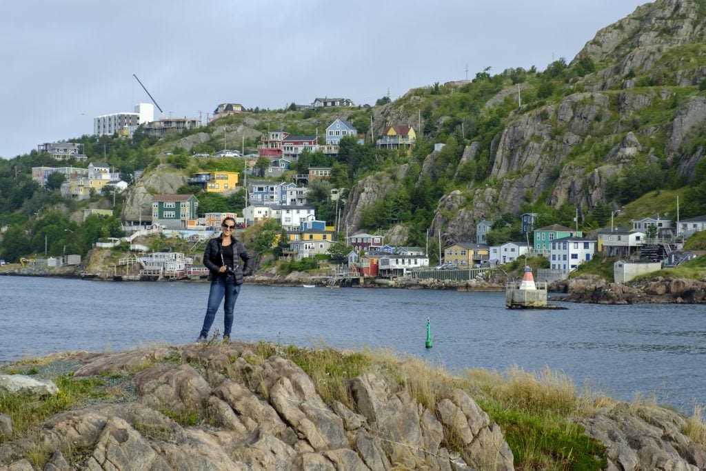 Kate stands on a cliff in Newfoundland in front of the bay and the colorful homes of St. John's behind her.