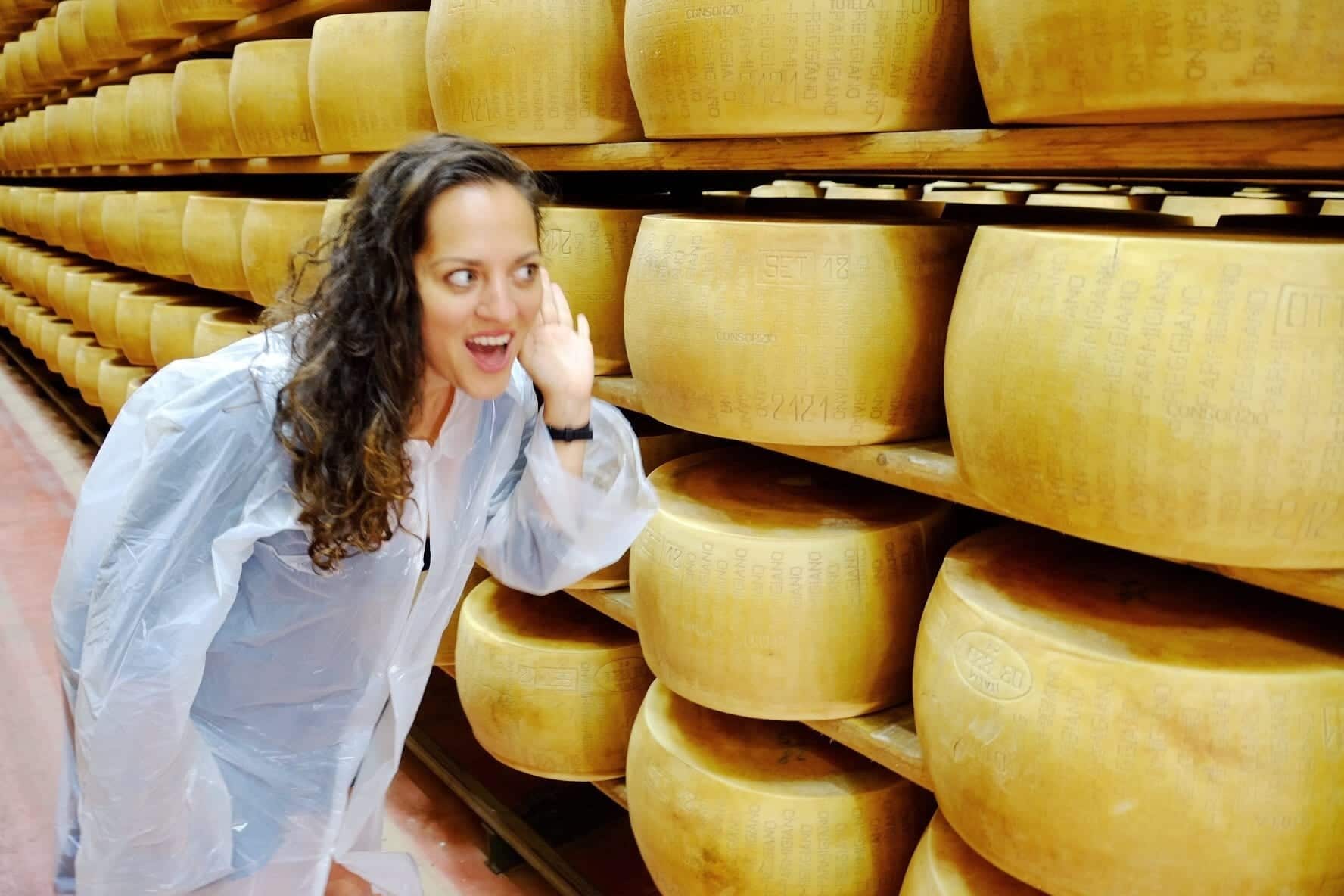 Kate stands in front of shelves of parmigiana reggiano wheels. She wears a protective robe and leans in with her hand to her ear, as if listening to the cheese.