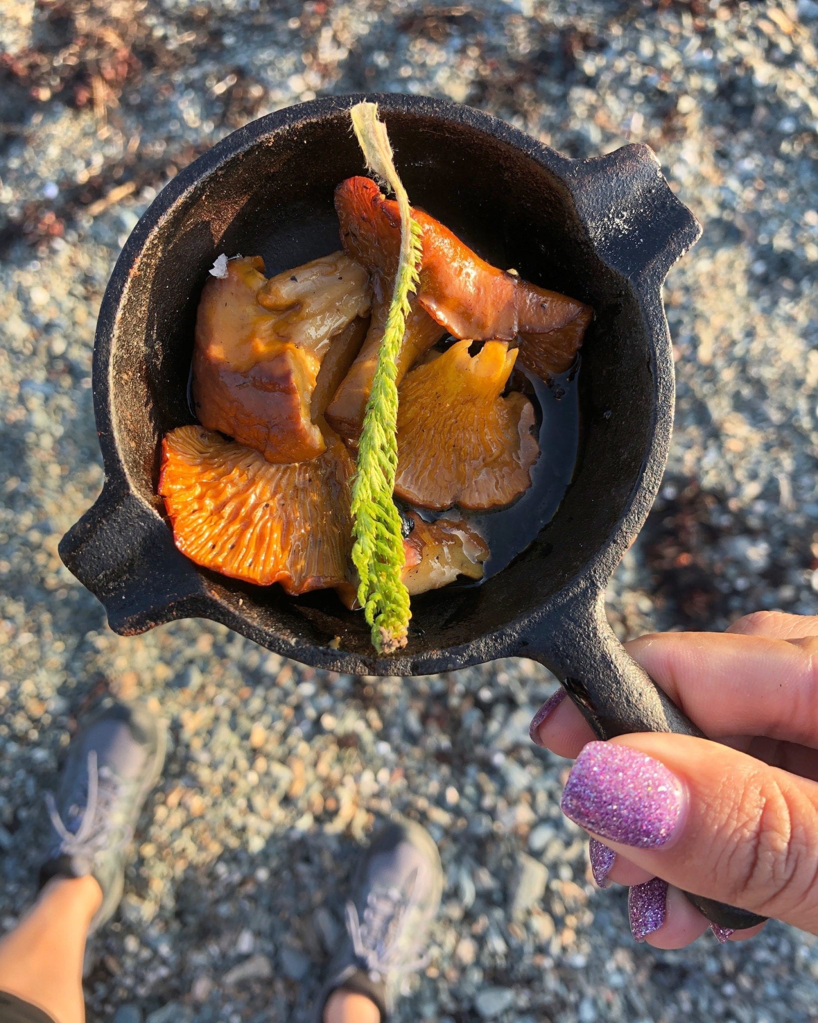 Kate holds a tiny cast iron skillet filled with chanterelle mushrooms and a spring of fresh green herbs. Beneath her you can see the sand and her purple hiking shoes,